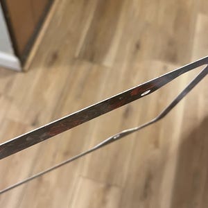Op de afbeelding: Een dun, rechthoekig metalen frame met een donker, gevlekt roodbruin en zwart patroon langs de randen. Het frame heeft kleine, ronde gaten met tussenpozen langs de dunne metalen rand, wat lijkt op een onderdeel van een groter geheel.