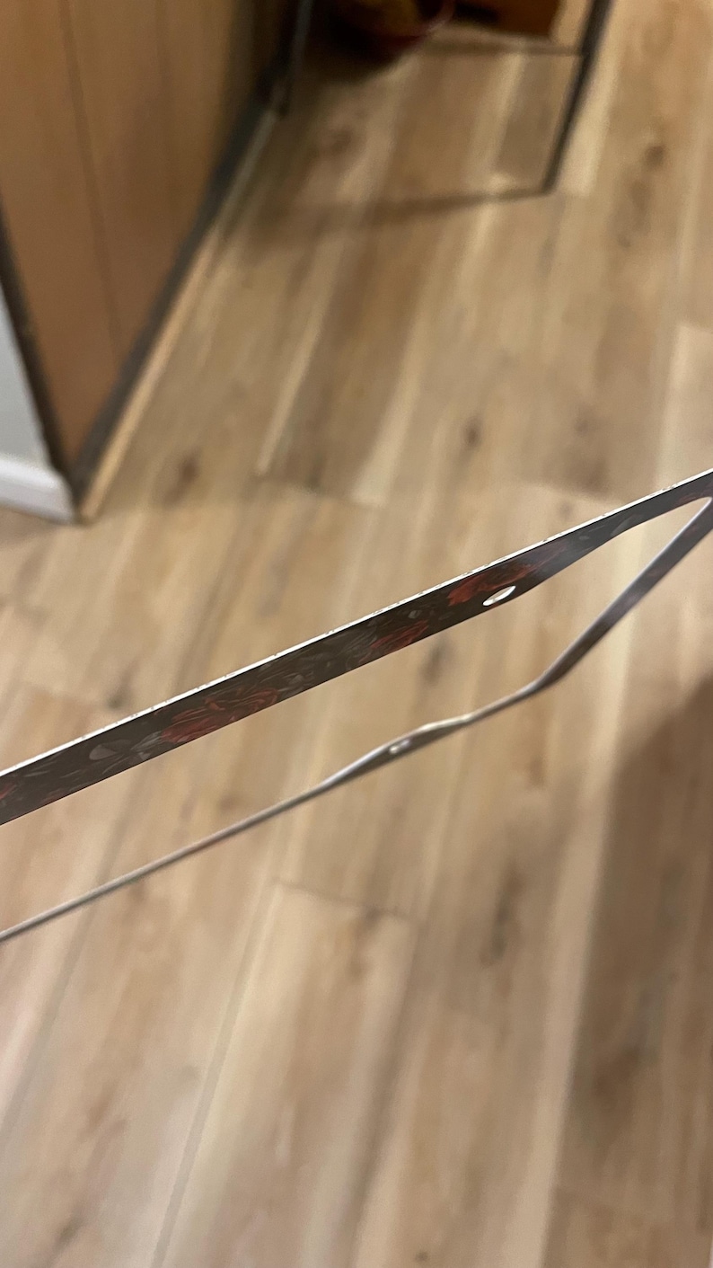 Op de afbeelding: Een dun, rechthoekig metalen frame met een donker, gevlekt roodbruin en zwart patroon langs de randen. Het frame heeft kleine, ronde gaten met tussenpozen langs de dunne metalen rand, wat lijkt op een onderdeel van een groter geheel.
