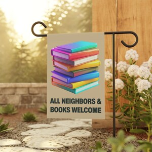 May include: A beige garden flag featuring a stack of colourful books. The books have vibrant covers in shades of blue, pink, red, yellow, and purple. The text "ALL NEIGHBOURS & BOOKS WELCOME" is printed in dark grey.