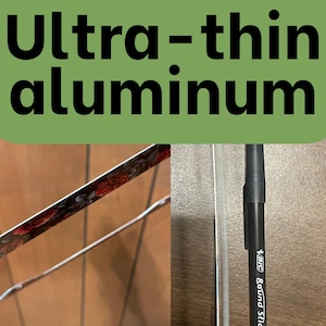 Op de afbeelding: Een ultra-dunne aluminium plaat met een donker bloemenpatroon met rode rozen aan &eacute;&eacute;n kant en een metalen rand. Een zwarte Bic Round Stic pen is verticaal naast de rand geplaatst om het dunne profiel van het materiaal te demonstreren.