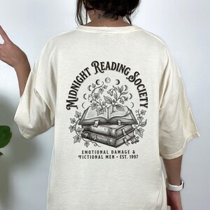 Puede incluir: Una camiseta color hueso con un gráfico negro en la espalda. El diseño muestra dos libros apilados con hiedra creciendo del superior, rodeados de fases lunares y el texto MIDNIGHT READING SOCIETY, EMOTIONAL DAMAGE & FICTIONAL MEN EST. 1997.
