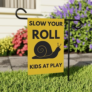 May include: A yellow garden flag with black text reading "SLOW YOUR ROLL" and "KIDS AT PLAY". A black snail graphic is centered on the flag. The flag is displayed on a black metal garden stake in a grassy garden.