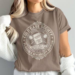 May include: A brown t-shirt featuring a circular graphic with the text COLD CASE SOCIETY, PLAUSIBLE DENIABILITY DIVISION, MUNICIPAL ARCHIVES, INVESTIGATION, TRUTH, JUSTICE, SPECIAL OPS, EST 1973, and a central illustration of a file and magnifying glasses.