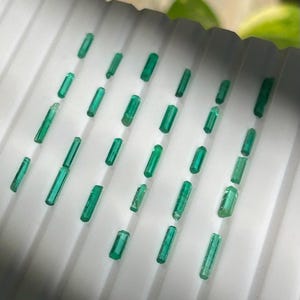 Op de afbeelding: Een verzameling kleine, levendige groene smaragdkristallen. De kristallen zijn rechthoekig en variëren enigszins in grootte en helderheid. Ze zijn gerangschikt op een wit, geribbeld oppervlak, wat een visueel aantrekkelijke weergave van natuurlijke edelstenen creëert.