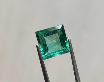 Puede incluir: Una esmeralda verde vibrante, de corte cuadrado, es sostenida por unas pinzas plateadas. La esmeralda tiene una superficie facetada, que refleja la luz y muestra su claridad. La gema es de un color verde intenso.