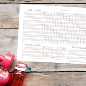 May include: A printable habit tracker with sections for daily, weekly, and monthly habits. The tracker is printed on white paper and has a light brown grid pattern. The tracker is laid out on a wooden surface with a red dumbbell, a red apple, and a red water bottle.
