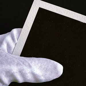 Puede incluir: Un guante de algod&oacute;n blanco sosteniendo un marco de fotos blanco vac&iacute;o con un fondo negro.