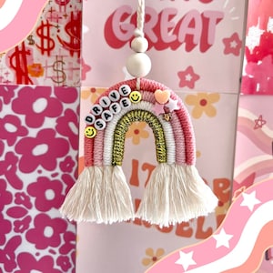 Puede incluir: Un adorno de coche de arcoíris de macramé hecho a mano en tonos rosa, blanco y dorado. El adorno tiene las palabras "DRIVE SAFE" deletreadas con cuentas de letras y una carita sonriente. Cuelga de un cordón blanco con cuentas de madera. El fondo presenta papel estampado rosa y blanco.