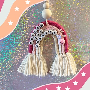 Puede incluir: Un tapiz de macramé arcoíris rosa y blanco con las palabras "Follow the Sparkle" en letras negras. El arcoíris tiene una borla blanca a cada lado y una cuenta de madera en la parte superior.