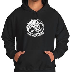 Pode incluir: Moletom preto com um design gráfico branco. O design apresenta um inseto estilizado dentro de um círculo, com as palavras "HOOK | TRAP | STRIKE" em texto branco. O moletom tem um bolso frontal e um capuz com cordão.