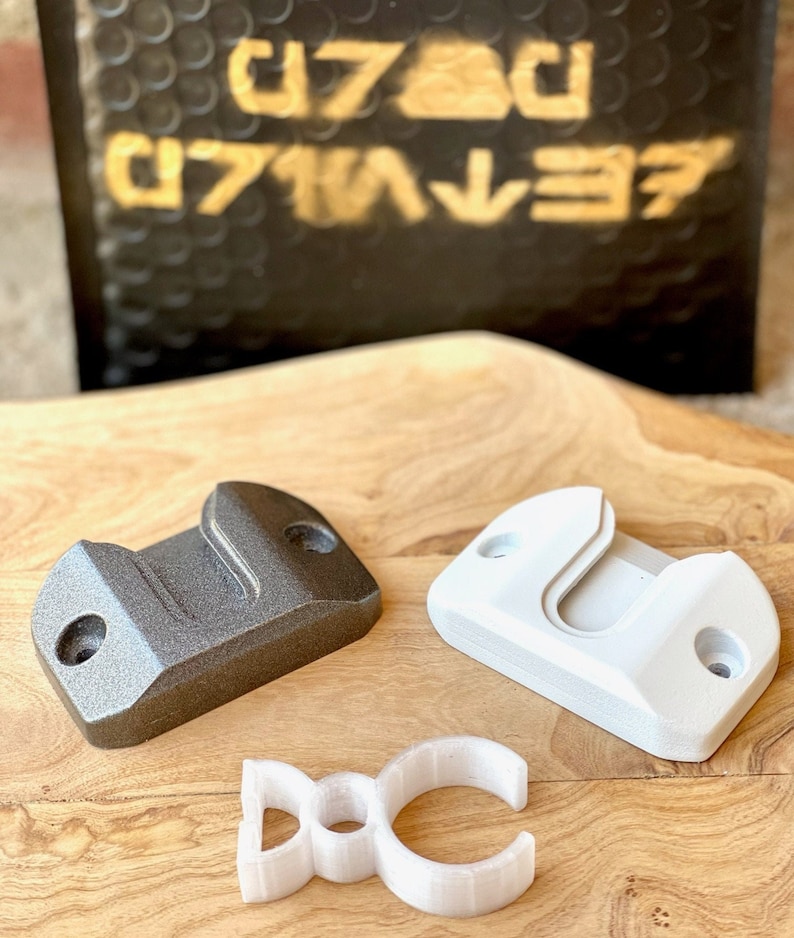 May include: Three 3D printed wall mounts in white and gray. The mounts are designed to hold a variety of objects, such as tools, keys, or other small items. The mounts are shown on a wooden surface.