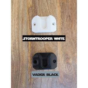 May include: Two 3D printed wall mounts for a lightsaber. One is white and labeled "STORMTROOPER WHITE". The other is black and labeled "VADER BLACK".