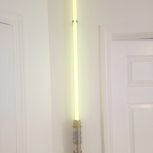 May include: A lightsaber wall mount with a silver and gold finish. The lightsaber is lit up with a bright green glow.