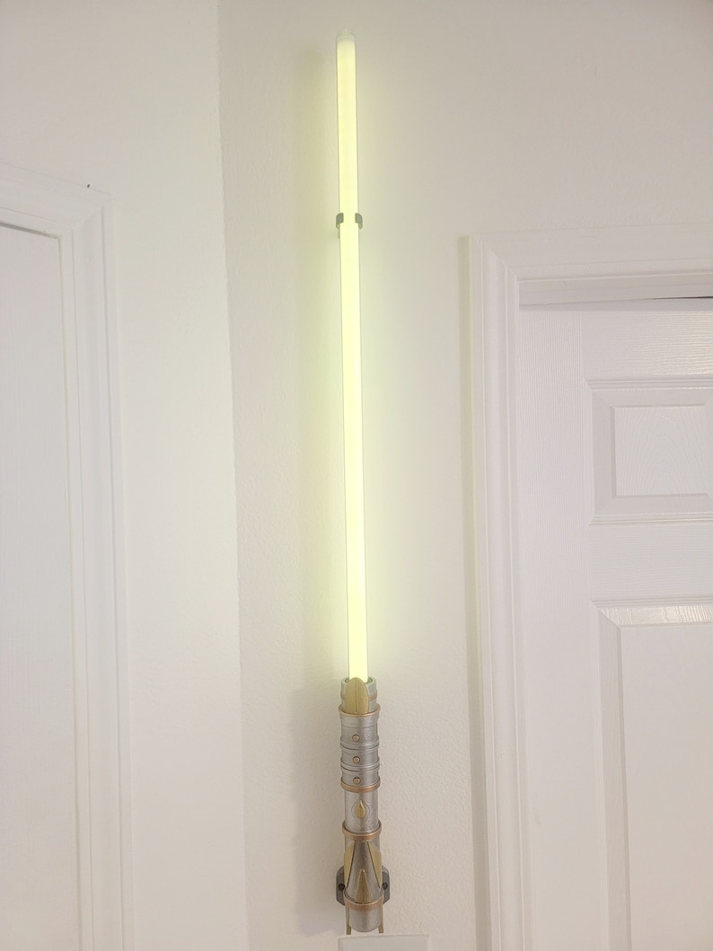 May include: A lightsaber wall mount with a silver and gold finish. The lightsaber is lit up with a bright green glow.