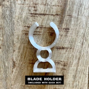 May include: A clear plastic blade holder, designed to hold a razor blade. The holder is shaped like a U with a rounded base. The text "BLADE HOLDER (INCLUDED WITH EACH SET)" is printed below the holder.