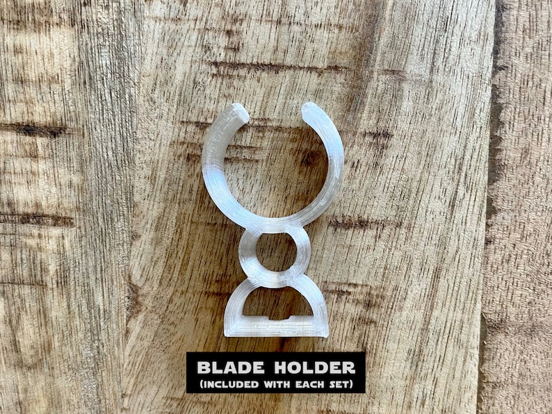 May include: A clear plastic blade holder, designed to hold a razor blade. The holder is shaped like a U with a rounded base. The text "BLADE HOLDER (INCLUDED WITH EACH SET)" is printed below the holder.