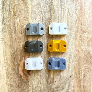 May include: Six wall mounts in different colors: gray, white, black, gold, white, and blue. The mounts are made of plastic and have a rectangular shape with a curved top.