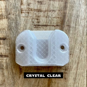May include: A clear plastic wall mount with two holes for screws. The mount has a U-shaped opening in the center.