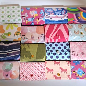 May include: A collection of small, decorative card holders with various patterned designs. The patterns include floral, geometric, and abstract motifs in a range of colors. Each holder has a button closure with a decorative gem.