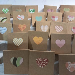 May include: A collection of small, square kraft paper cards, each adorned with a heart-shaped embellishment. The hearts vary in color and pattern, including stripes, polka dots, and solid hues. The cards are arranged closely together.