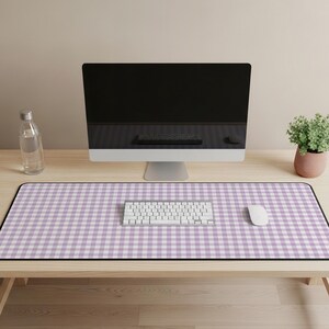 Pode incluir: Uma configuração de mesa com um tapete de mesa retangular grande com um padrão xadrez roxo e branco. Um teclado e um mouse brancos estão no tapete. Um monitor de computador, uma garrafa de água e uma planta em vaso também estão na mesa de madeira.