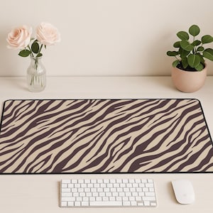 May include: A long rectangular desk mat with a zebra stripe pattern in brown and tan. The mat has a black border and is placed on a white desk. A white keyboard and mouse are in the foreground.