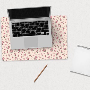 K&ouml;nnte beinhalten: Ein wei&szlig;er Laptop mit schwarzem Bildschirm liegt auf einer rosa-wei&szlig;en Blumen-Schreibtischunterlage. Ein wei&szlig;es Spiralnotizbuch und ein brauner Bleistift liegen auf einer wei&szlig;en Oberfl&auml;che.