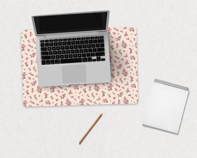 K&ouml;nnte beinhalten: Ein wei&szlig;er Laptop mit schwarzem Bildschirm liegt auf einer rosa-wei&szlig;en Blumen-Schreibtischunterlage. Ein wei&szlig;es Spiralnotizbuch und ein brauner Bleistift liegen auf einer wei&szlig;en Oberfl&auml;che.