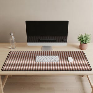 May include: A brown and white gingham pattern desk mat with a black border. A white keyboard and mouse are on the mat. A computer monitor, a small potted plant, and a water bottle are also on the wooden desk.