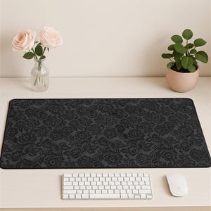 May include: A black rectangular desk mat with a floral lace pattern. The mat is on a white desk, with a white keyboard and mouse in front. A vase of pink roses and a potted plant are also on the desk.