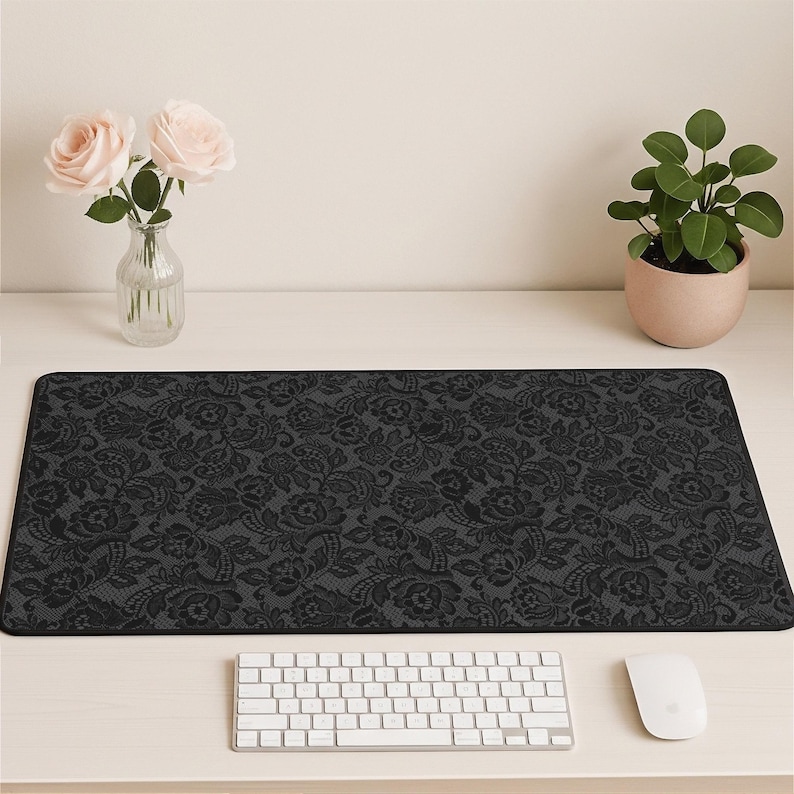 May include: A black rectangular desk mat with a floral lace pattern. The mat is on a white desk, with a white keyboard and mouse in front. A vase of pink roses and a potted plant are also on the desk.