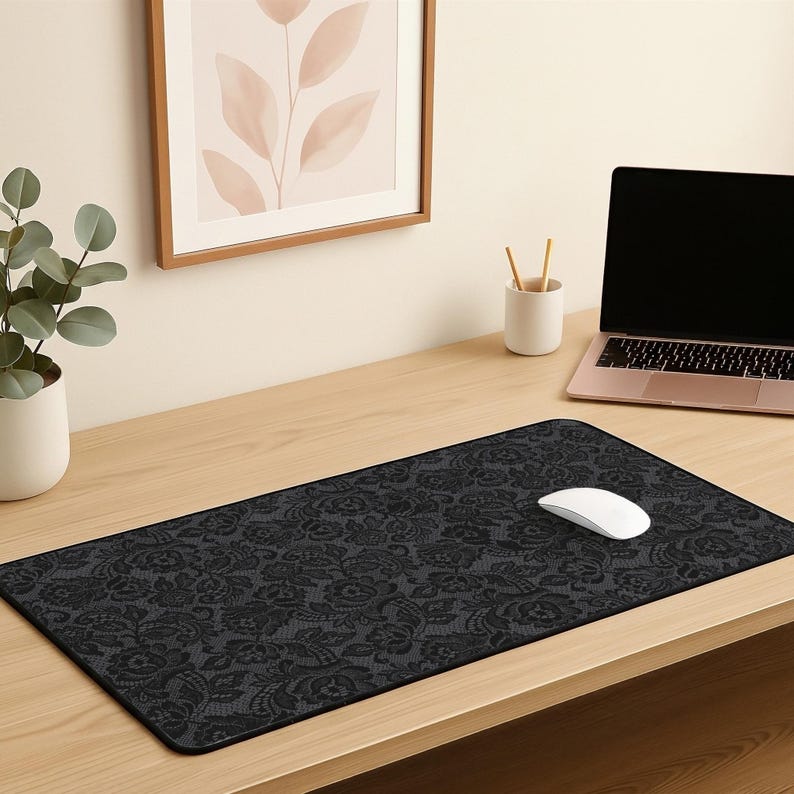 May include: A black rectangular mouse pad with a floral lace pattern. A white computer mouse rests on the pad. The pad is on a wooden desk, with a laptop and a framed picture in the background.
