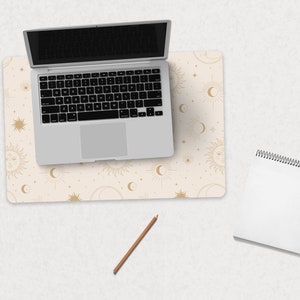 May include: A beige desk pad with a gold celestial pattern featuring suns, moons, and stars. A silver laptop computer is on top of the desk pad. A white spiral bound notebook and a brown pencil are on the desk.