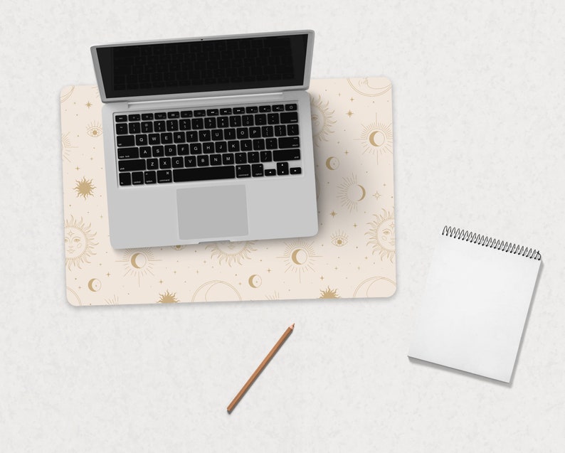 May include: A beige desk pad with a gold celestial pattern featuring suns, moons, and stars. A silver laptop computer is on top of the desk pad. A white spiral bound notebook and a brown pencil are on the desk.