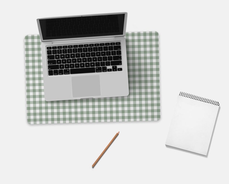 Pode incluir: Um computador port&aacute;til branco com um teclado preto est&aacute; em uma esteira de mesa quadriculada verde e branca. Um caderno branco em espiral e um l&aacute;pis marrom est&atilde;o na superf&iacute;cie branca.