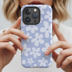 Pode incluir: Uma capa de telefone azul claro com um padrão floral branco. A capa tem um acabamento fosco e um protetor de lente de câmera preto. A capa do telefone é segurada por duas mãos.
