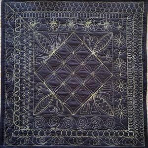 May include: A dark blue quilted fabric with a geometric pattern stitched in a light yellow thread. The fabric has a border of various decorative stitches.