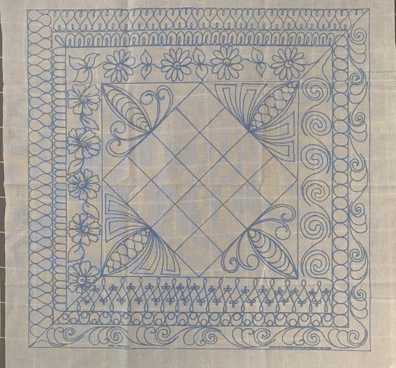 May include: A white fabric with a blue design of a square with a diamond pattern inside. The square is surrounded by a border of floral and swirl designs. The fabric is marked with the text "www.stitcherydesigns.com/free.trial"