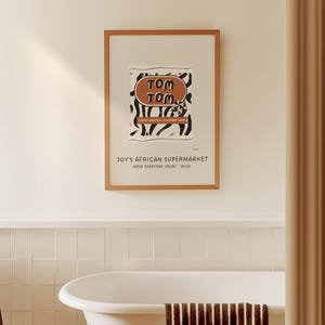 May include: A framed poster with a brown wooden frame hangs on a white wall. The poster features the text "TOM TOM" in a brown oval, with black and white abstract shapes. Below, it reads "JOY'S AFRICAN SUPERMARKET" and the hours of operation.