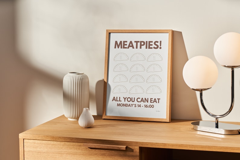 K&ouml;nnte beinhalten: Ein gerahmter Druck mit dem Text "MEAT PIES! ALL YOU CAN EAT MONDAY'S 14 - 16:00" und einer Grafik von 12 Fleischpasteten. Der Druck befindet sich auf einem Holztisch mit einer wei&szlig;en Vase und einer kleinen wei&szlig;en Vase.