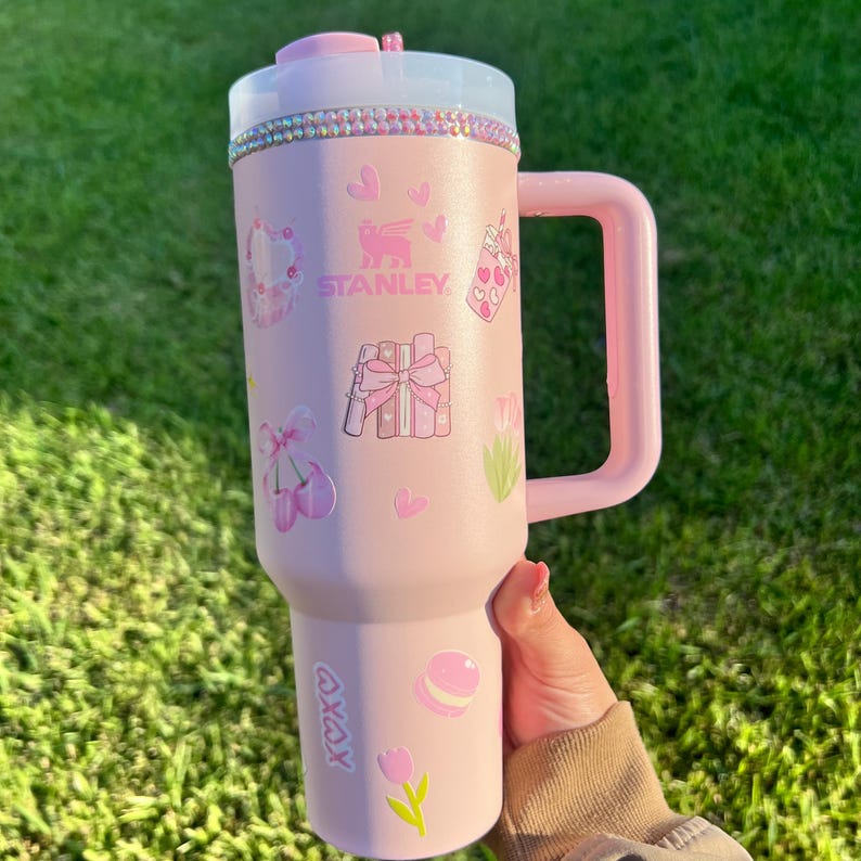 Puede incluir: Un vaso Stanley rosa con asa, decorado con pegatinas de corazones, lazos y macarons. La tapa es rosa y transparente, con una banda de pedrer&iacute;a. El vaso tiene el logotipo de Stanley y el texto "XOXO".