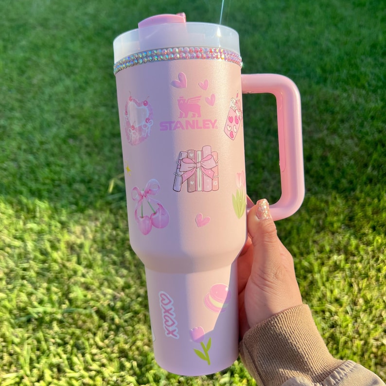 Puede incluir: Un vaso Stanley rosa con asa, decorado con corazones, regalos y zapatillas de ballet. El borde superior est&aacute; adornado con pedrer&iacute;a. El vaso tiene una tapa transparente y el logotipo de Stanley es visible. Sobre un fondo de hierba verde.