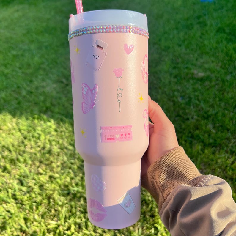 Puede incluir: Un vaso rosa con tapa blanca y pajita rosa. El vaso est&aacute; decorado con pegatinas rosas de una mariposa, un frasco de perfume, corazones y la palabra "kiss". El borde del vaso est&aacute; adornado con pedrer&iacute;a.