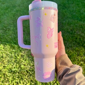 Puede incluir: Un vaso t&eacute;rmico rosa con asa, decorado con mariposas rosas, lazos y gr&aacute;ficos de &aacute;ngeles. El borde superior est&aacute; adornado con pedrer&iacute;a. El vaso tiene una tapa transparente y un asa rosa. Sobre un fondo de hierba verde.