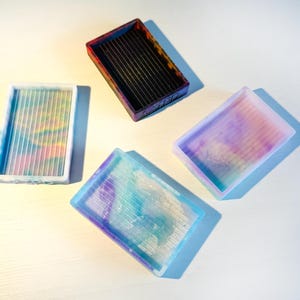 Pode incluir: Quatro tabuleiros de classificação retangulares com superfícies ranhuradas. Um tabuleiro é preto com bordas laranja, enquanto os outros três apresentam padrões de cores pastel translúcidas e rodopiantes em azul, rosa e branco. Cada tabuleiro tem as letras TPD gravadas.