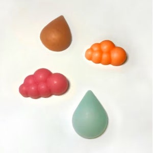 Puede incluir: Cuatro barras de jabón hechas a mano en forma de nubes y gotas de lluvia. Los jabones son en tonos de naranja, rosa y azul claro.