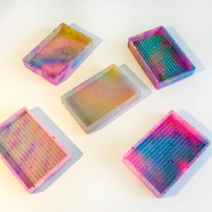 Puede incluir: Cinco bandejas rectangulares de resina con interior acanalado. Las bandejas son de varios colores, incluyendo rosa, azul, amarillo y morado, con un efecto tie-dye moteado. Las bandejas están diseñadas para manualidades.
