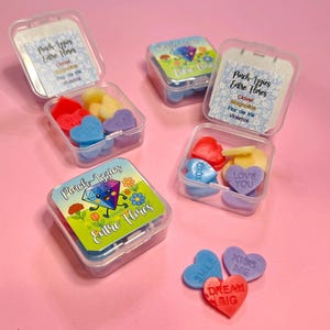 Puede incluir: Pequeños recipientes de plástico transparente llenos de ceras perfumadas en forma de corazón de varios colores. Los recipientes tienen etiquetas con texto y diseños florales. También se ven ceras en forma de corazón sueltas con frases.
