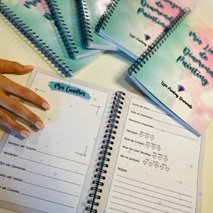 Puede incluir: Cuadernos encuadernados en espiral con una cubierta pastel, con el texto "Mis Lico de Diamond Painting". El cuaderno abierto muestra páginas rayadas para el seguimiento de proyectos de pintura de diamantes. Los cuadernos están apilados sobre una superficie blanca.