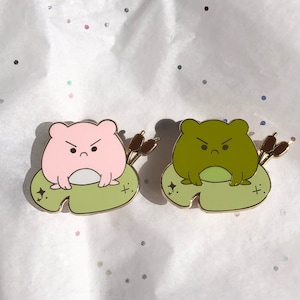 May include: Two enamel pins featuring cartoon frogs with angry expressions. The frogs are sitting on lily pads with reeds behind them. One frog is pink and the other is green.